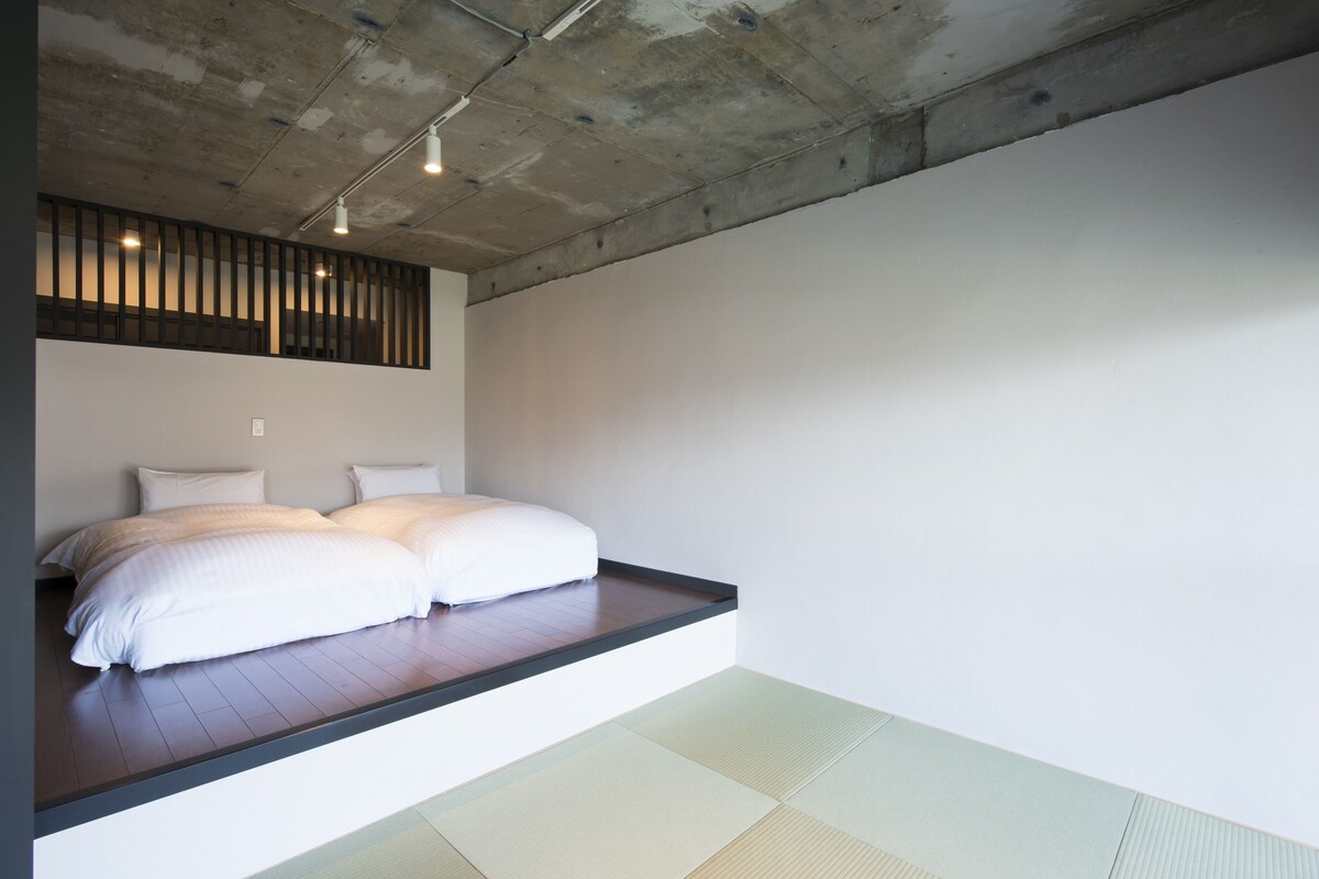 A minimalist bedroom with a low platform bed, tatami flooring, and a concrete ceiling.