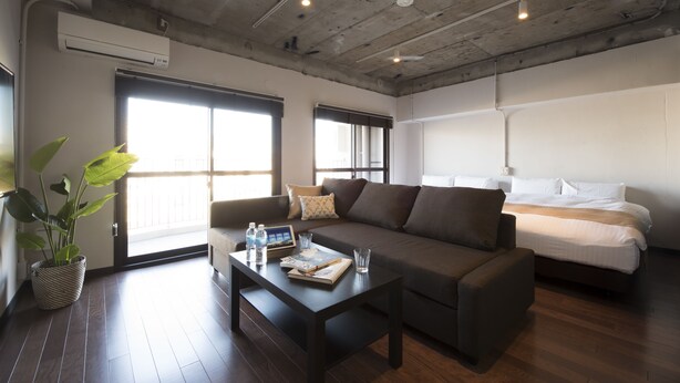 A modern hotel room with a large bed, a sofa, a coffee table with books and a water bottle, and a potted plant. - A Good Day Fukuoka Riverside (Fukuoka)