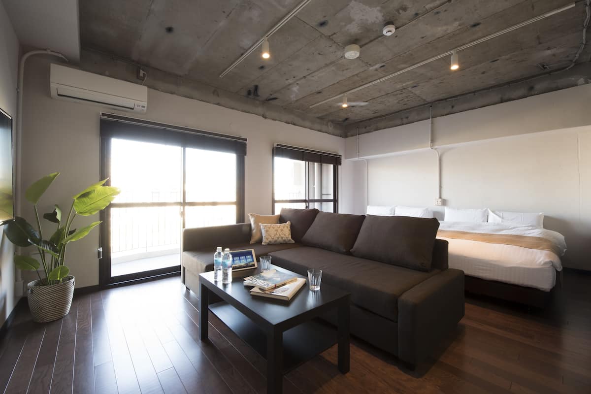 A modern hotel room with a large bed, a sofa, a coffee table with books and a water bottle, and a potted plant.