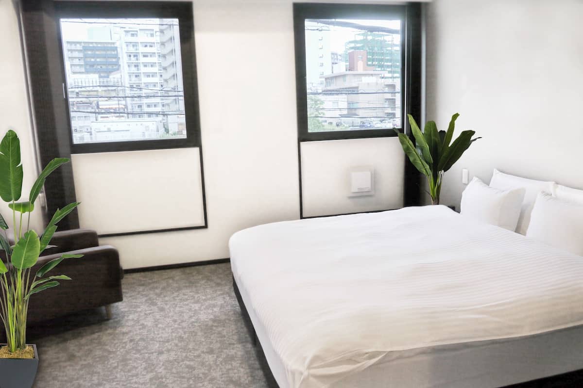 A modern hotel room with a large bed, two windows with city views, a small seating area, and two potted plants.