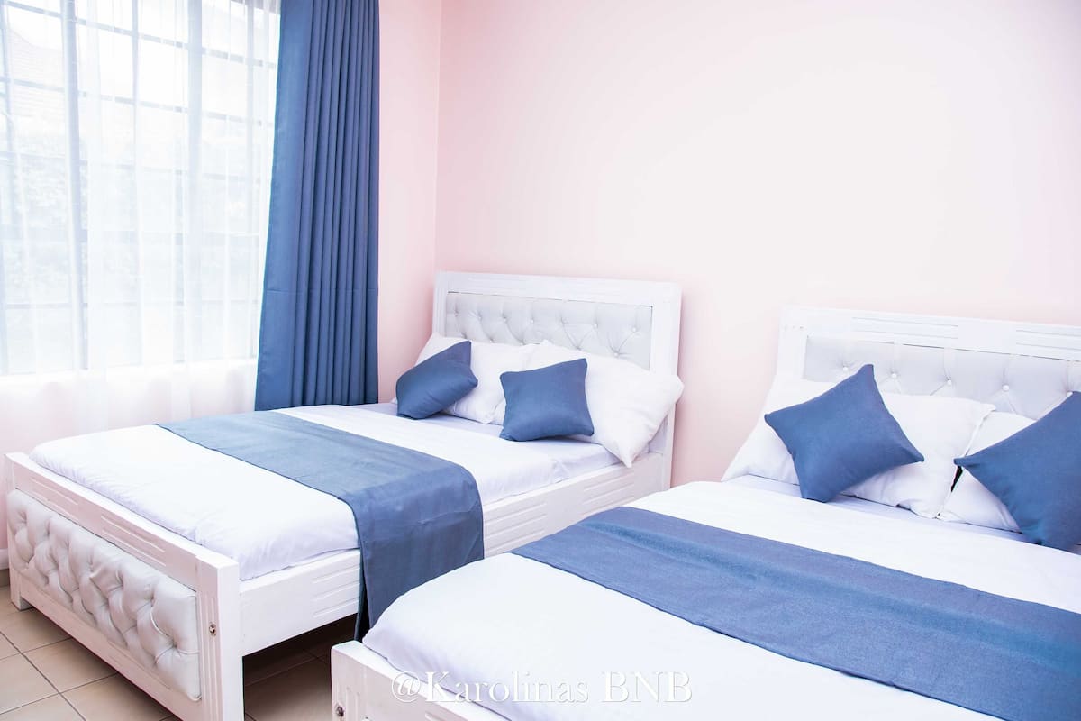 Two twin beds with white headboards and blue bedding in a room with a pink wall and a window with blue curtains.