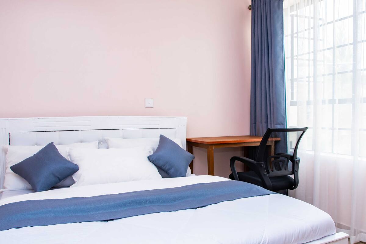 A neatly made bed with pillows, a small wooden table, a chair, and a window with curtains.