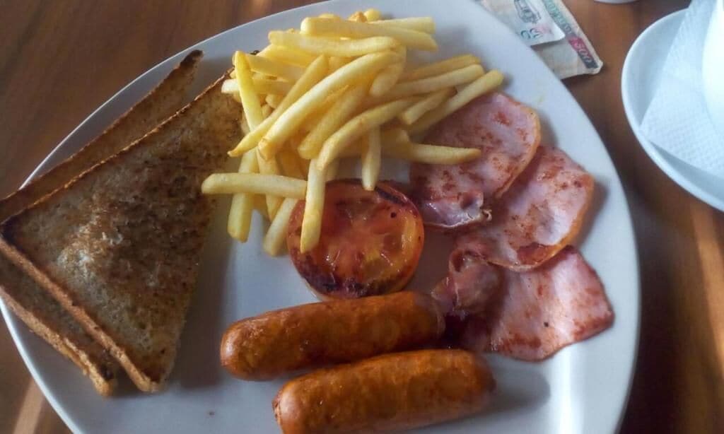 A plate with toast, French fries, bacon, sausages, and a tomato.