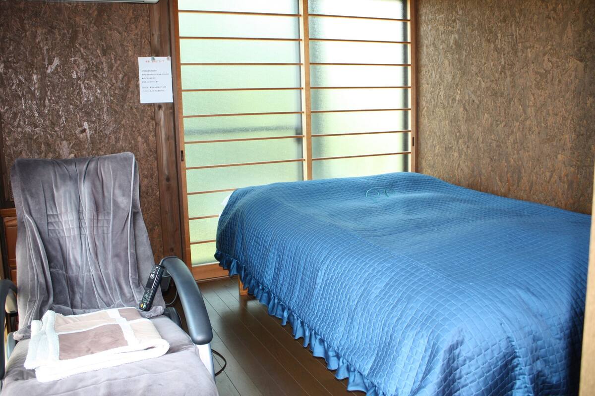 Ein kleines Zimmer mit einem Bett, einem Sessel und einem Fenster mit Jalousien.