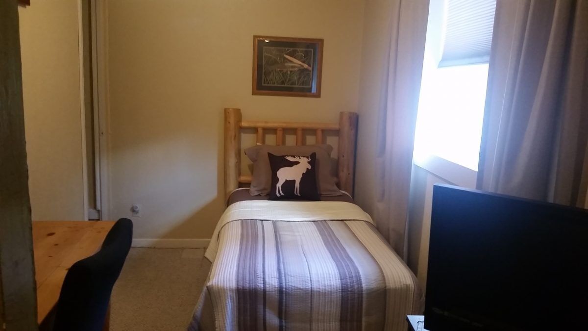 Ein kleines, gemütliches Zimmer mit einem Einzelbett, einem hölzernen Kopfteil, einem gerahmten Bild an der Wand und einem Fenster mit Vorhängen.