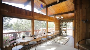A wooden interior with multiple round tables and chairs, large windows offering outdoor views, and a ceiling fan. - Jewel Lake Bed and Breakfast (Anchorage)