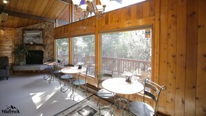 A wooden interior with a stone fireplace, a dining area with round tables and metal chairs, and large windows offering a view of trees. - Jewel Lake Bed and Breakfast (Anchorage)