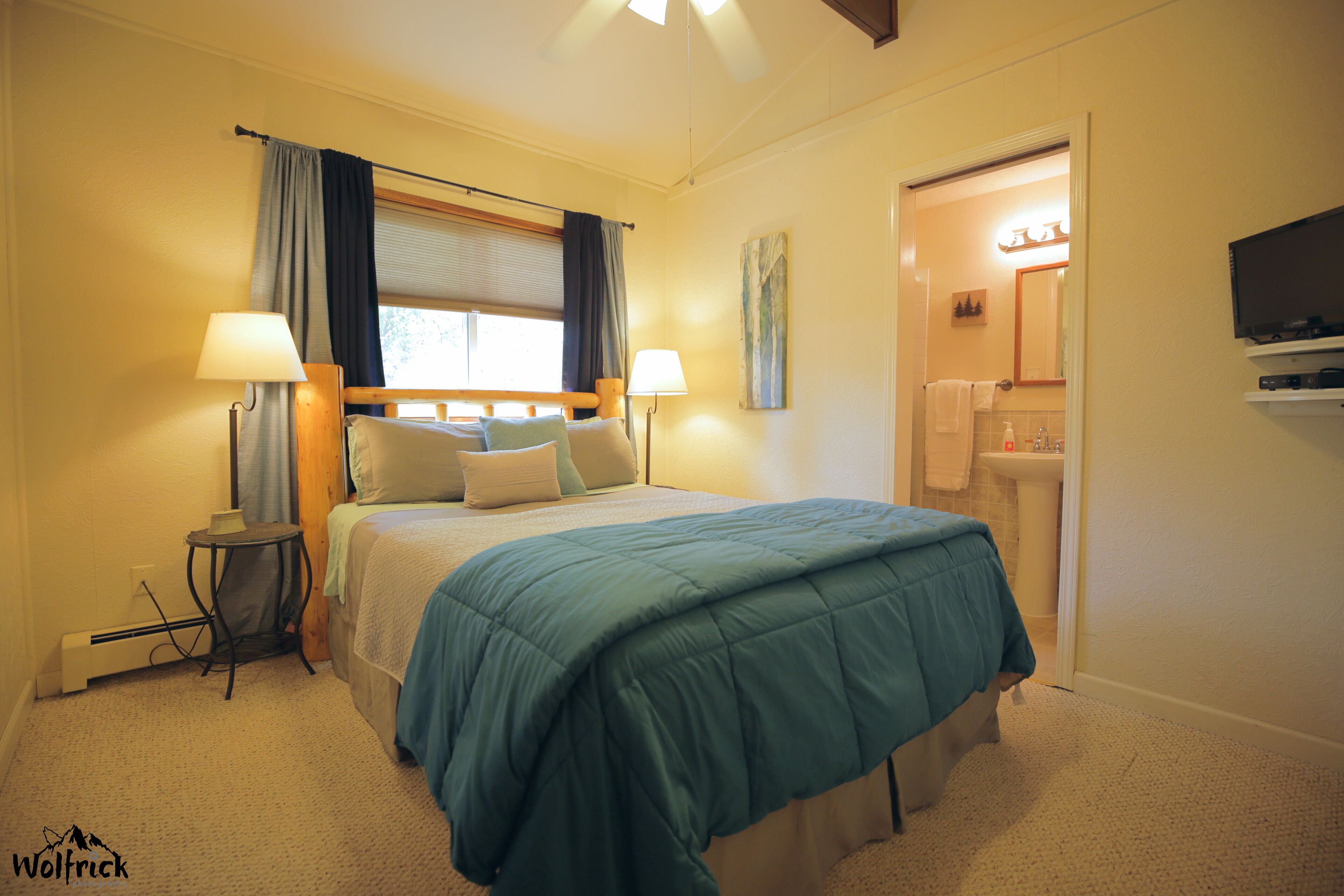 A hotel room with a bed, two bedside lamps, a window with curtains, a wall-mounted TV, and a bathroom visible through an open door.