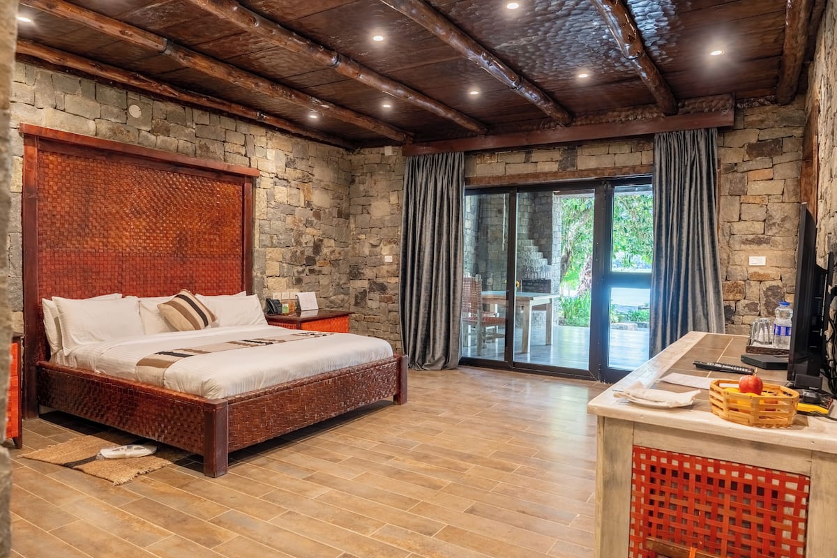 Une chambre avec un mur en pierre, un plafond en bois, un lit, une table de chevet, un bureau avec un panier, et une porte-fenêtre coulissante donnant sur l’extérieur.