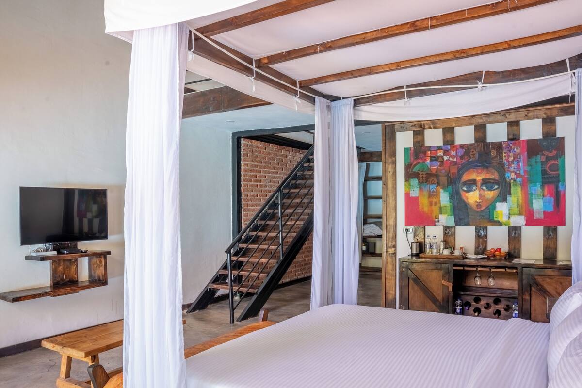 Une chambre à coucher avec un lit, une télévision, un banc en bois, un escalier et un tableau coloré.