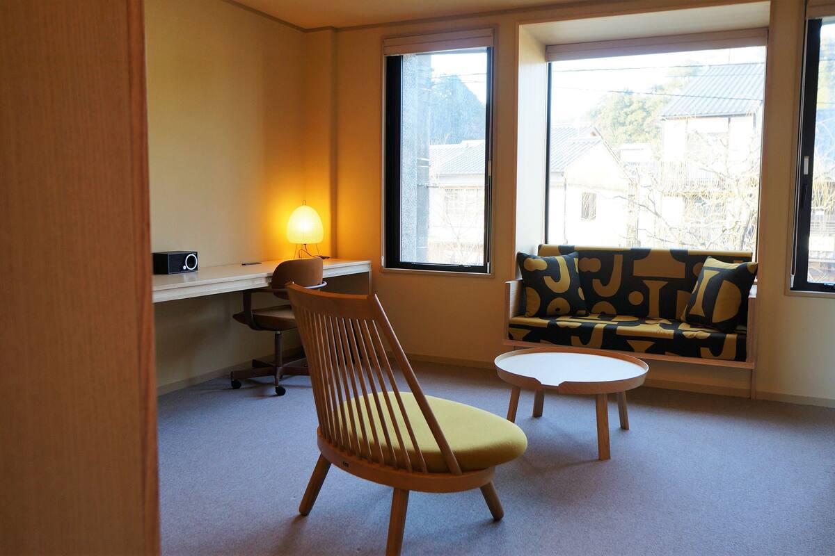 Ein Zimmer mit einem Holzstuhl, einer Couch mit gemusterten Kissen, einem Schreibtisch mit Lampe und großen Fenstern, die einen Blick auf Gebäude und Bäume bieten.