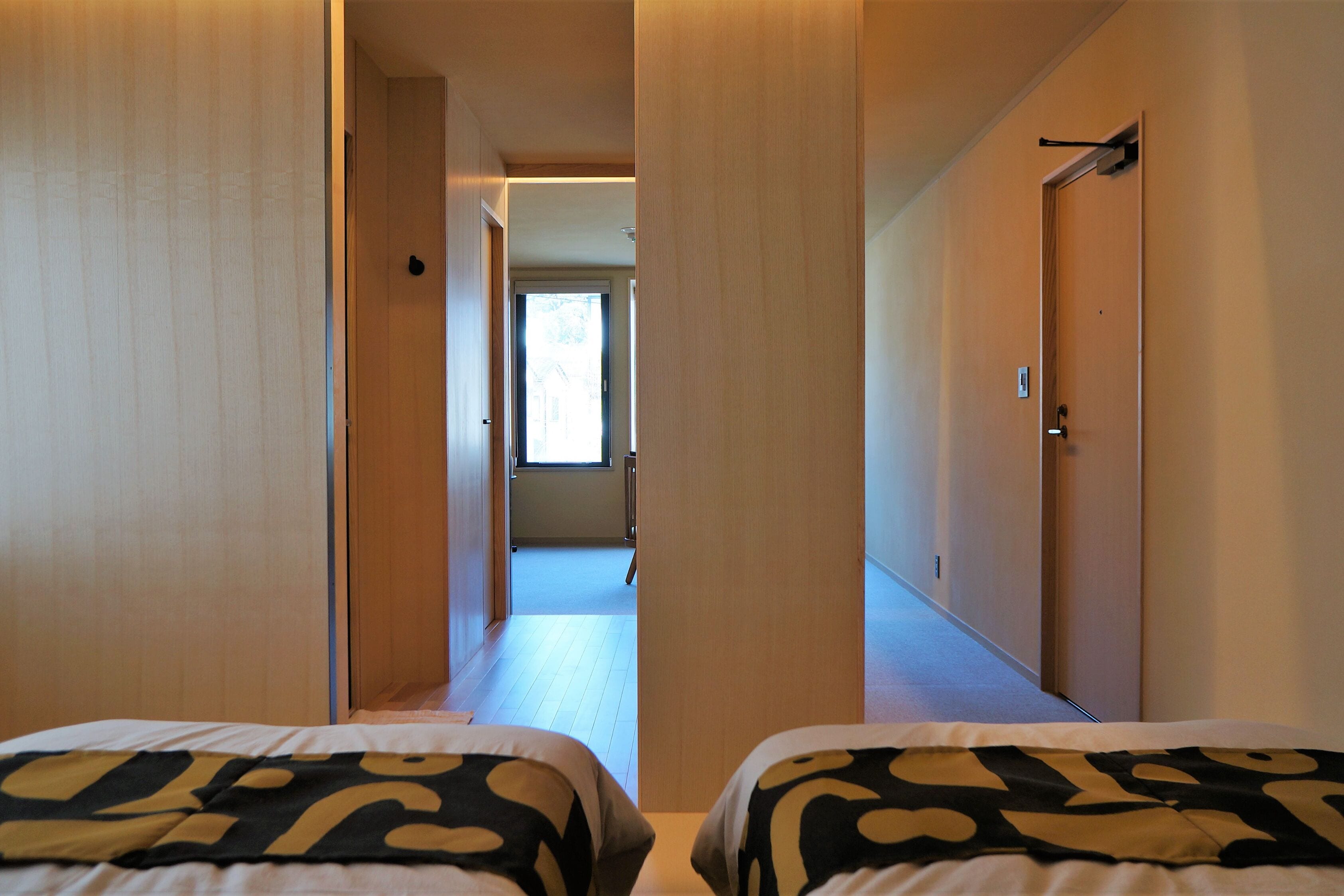 A hotel room with two beds, a wooden door, and a hallway leading to a bright room.