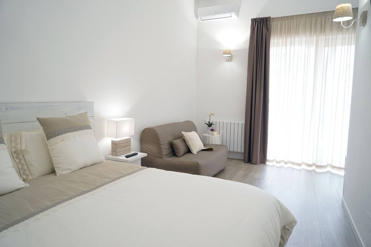 Ein Hotelzimmer mit Bett, Sofa, Nachttisch, Lampe, einem fenster mit Vorhängen und einer Klimaanlage.