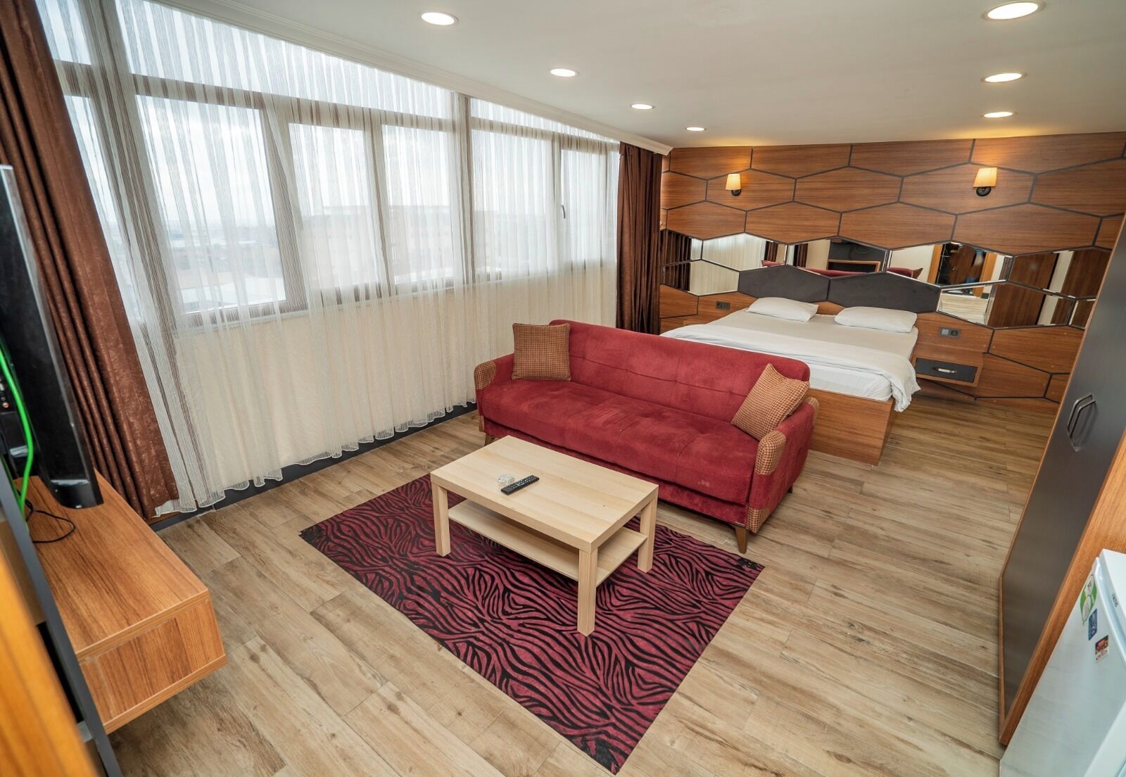 A hotel room with a red sofa, a wooden coffee table, a zebra-patterned rug, a bed with a wooden headboard, and a large window with sheer curtains.