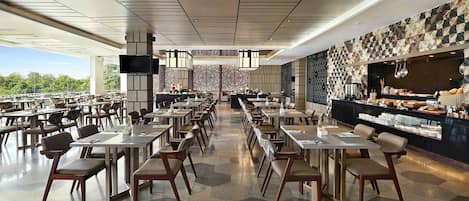 A spacious restaurant with multiple tables and chairs, a large counter with food display, and a television screen.