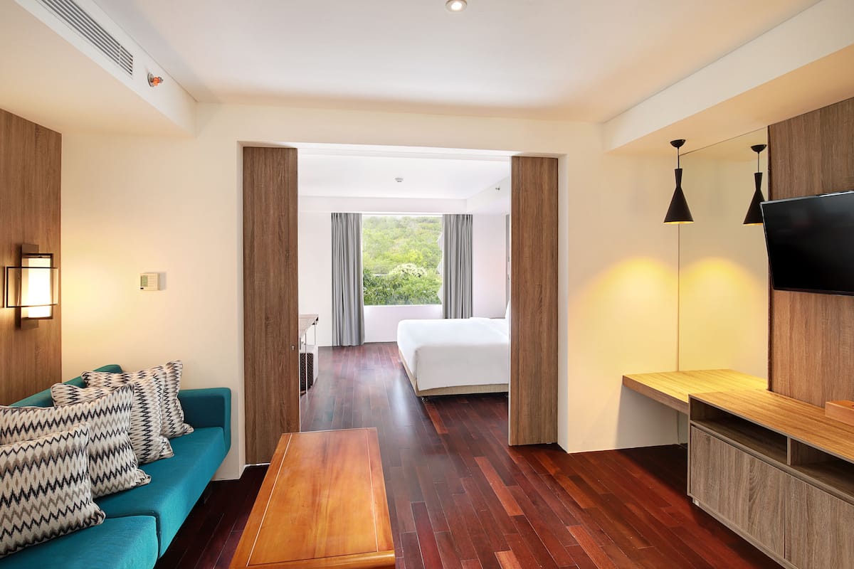 A modern hotel room with a wooden floor, a teal sofa with patterned cushions, a wooden bench, a bed with grey curtains, a TV mounted on the wall, and a view of greenery through the window.