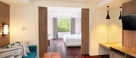 Una moderna camera d'albergo con pavimento in legno, un divano color petrolio con cuscini decorati, una panca in legno, un letto con tende grigie, una TV a parete e vista sul verde dalla finestra.