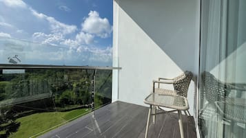 Un balcón con una silla de mimbre, baranda de vidrio y vistas a la vegetación y un cielo despejado.
