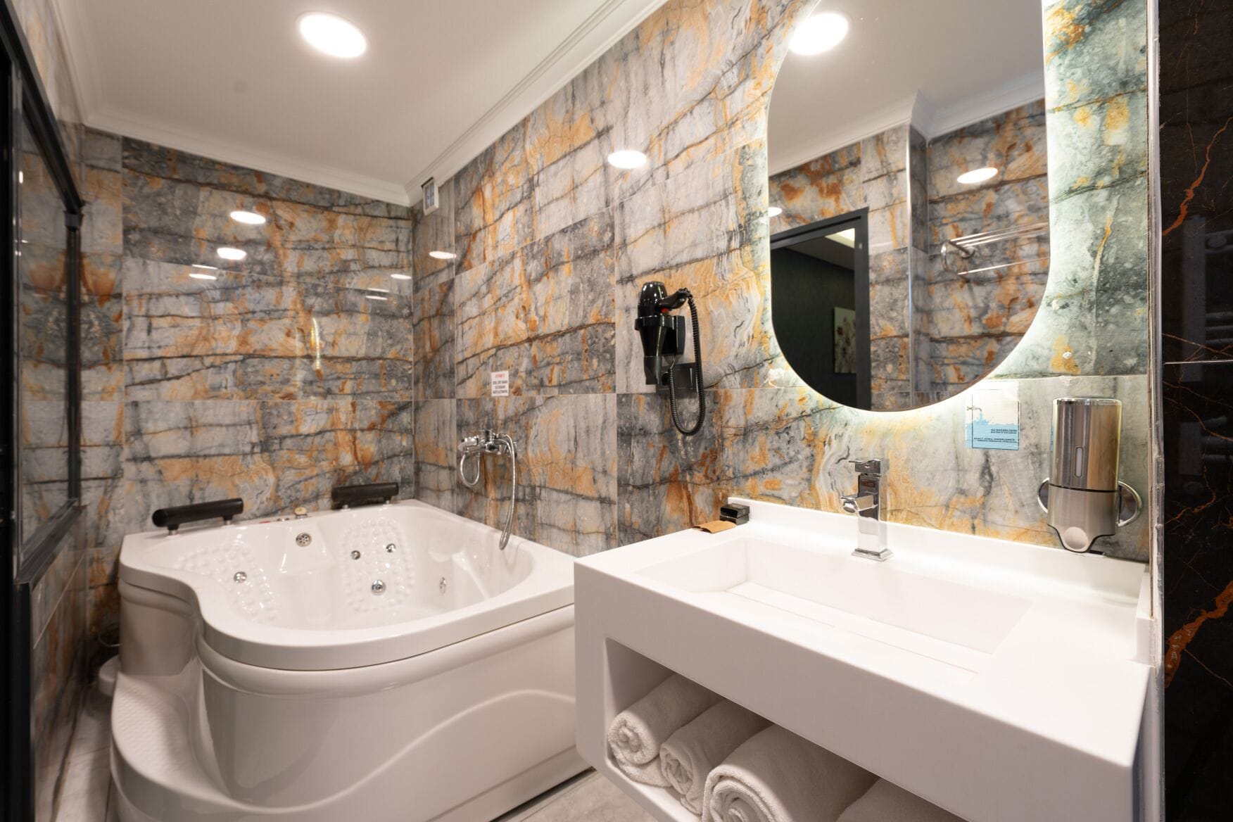 A modern bathroom with a marble wall, a large bathtub, a vanity with a mirror, and a towel rack.