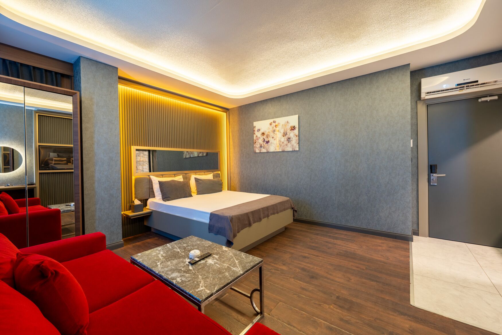 A modern hotel room with a bed, red sofa, bedside table, and a wall-mounted TV.