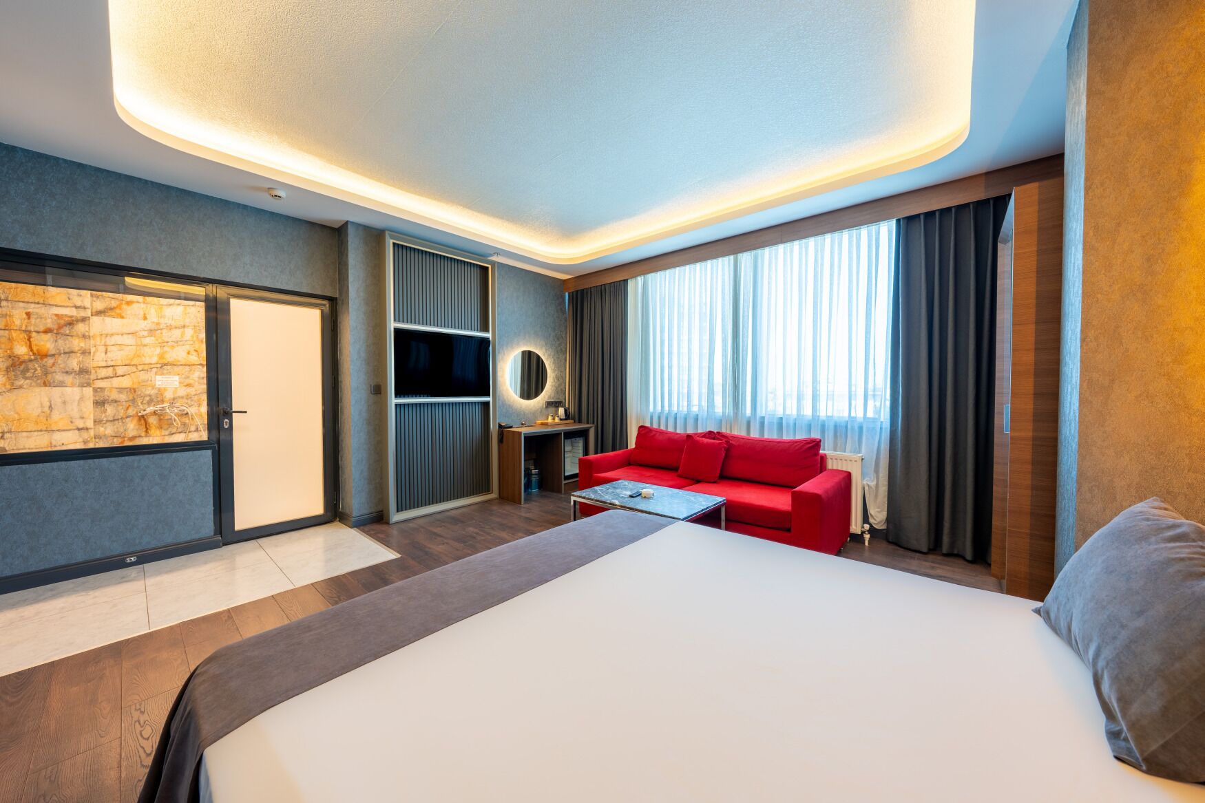 A modern hotel room with a large bed, a red sofa, a desk with a lamp, and a large window with curtains.