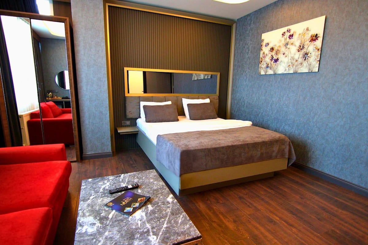 A hotel room with a bed, a red sofa, a small table with a remote control, and a painting on the wall.