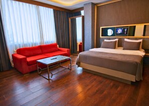 A hotel room with a red sofa, a bed with a grey duvet, wooden flooring, and a television on the wall. -  216 Eagle Palace Suite (Istanbul)