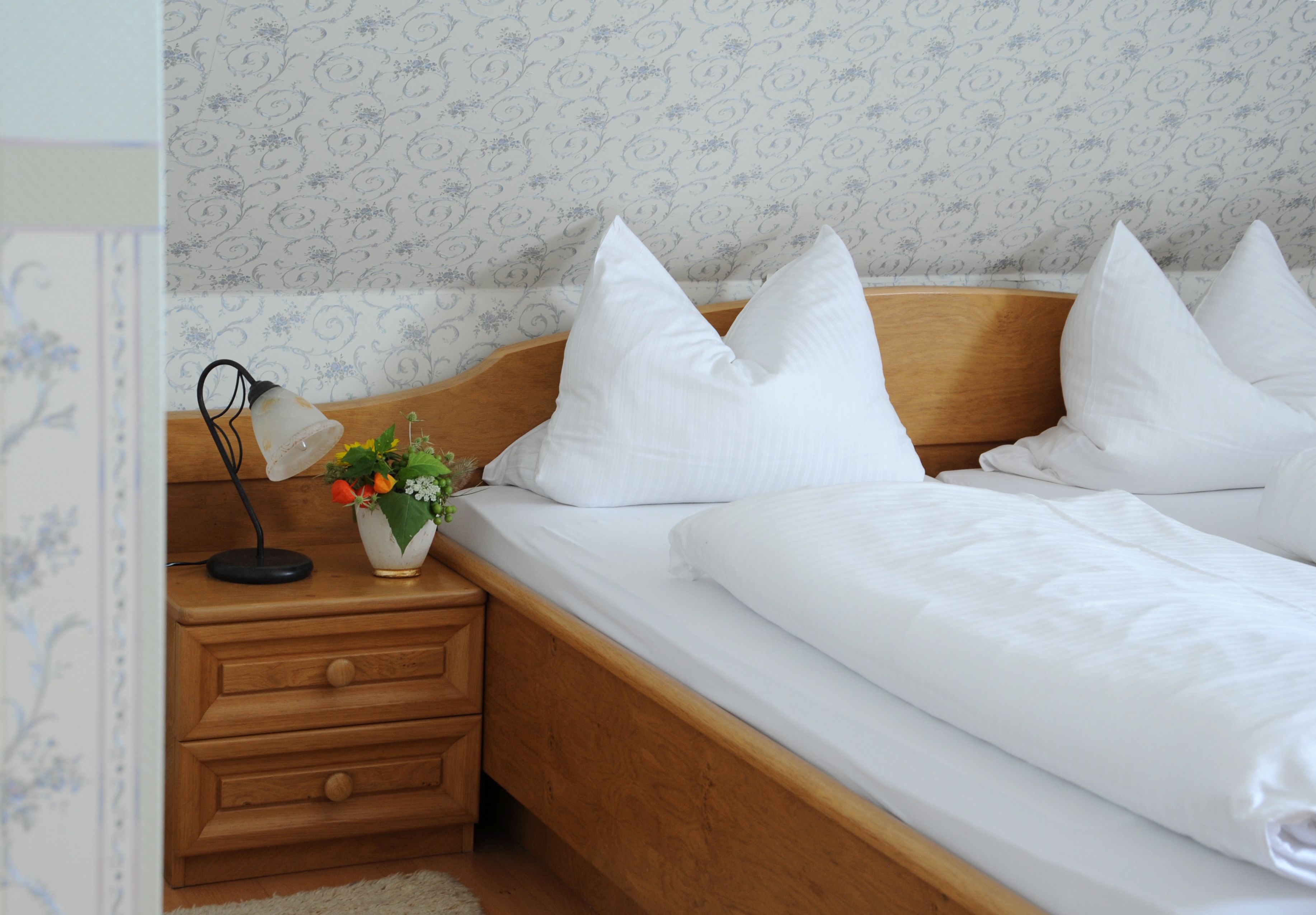 A neatly made bed with white bedding and pillows, a bedside table with a lamp and a vase of flowers, and a wooden nightstand.
