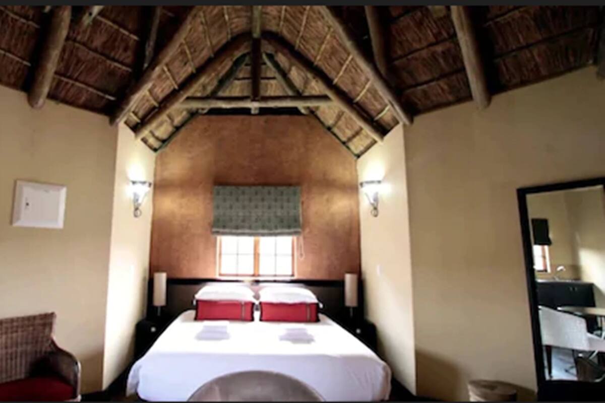 A room with a high ceiling, a bed with red pillows, a chair, a nightstand, and a mirror.