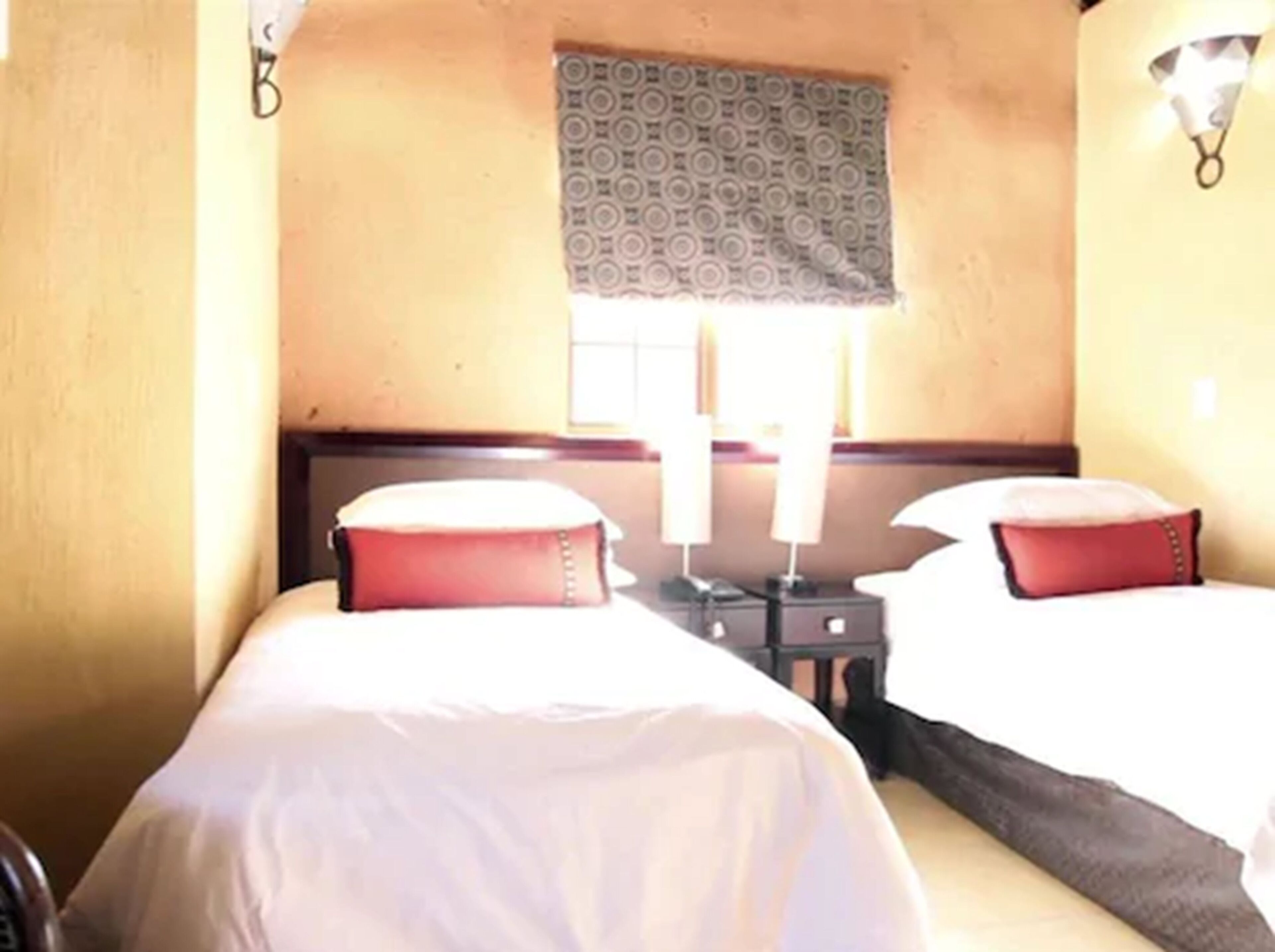 Une chambre d’hôtel avec deux lits, une fenêtre avec des rideaux à motifs, des lampes de chevet et une petite table placée entre les lits.