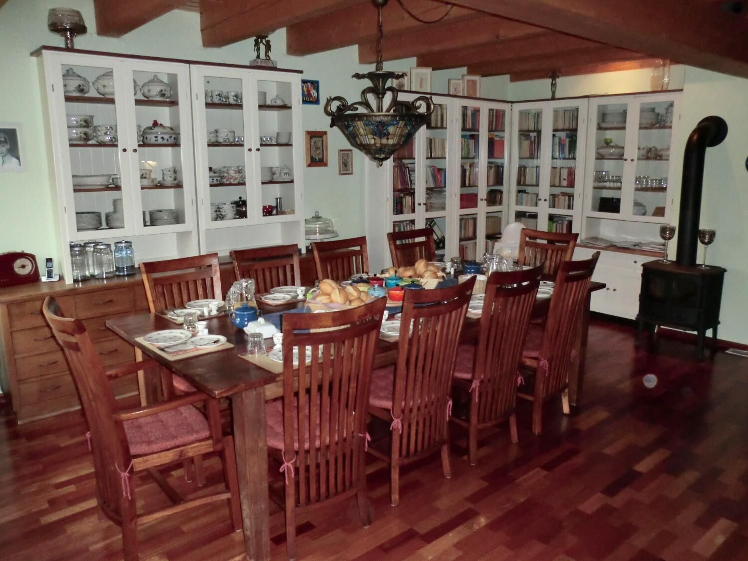 Comedor con una mesa de madera dispuesta para seis personas, rodeada de sillas de madera. En las paredes hay armarios con puertas de vidrio, y una estufa negra en la esquina.