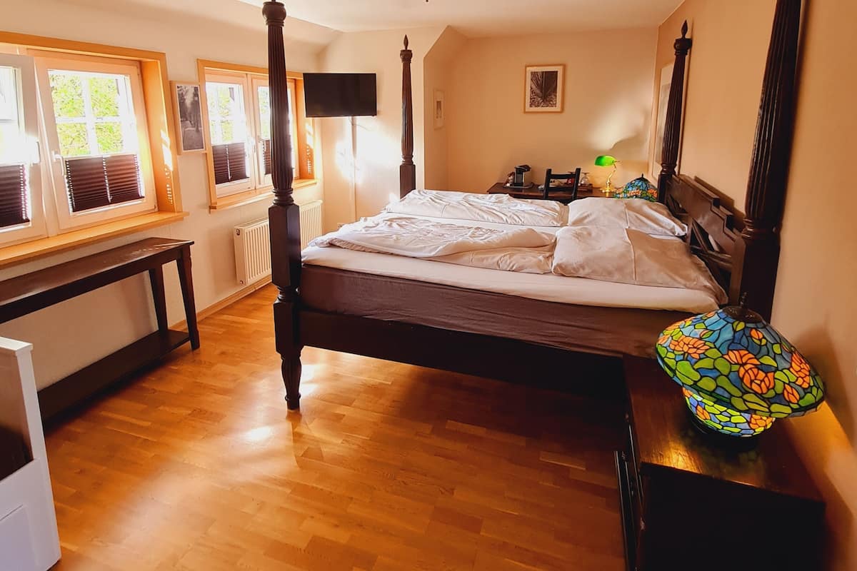 A bedroom with a four-poster bed, a desk, a chair, a lamp, and a window with blinds.