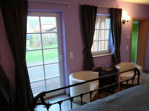 Una habitación con cama, una mesita con bandeja, dos sillas y un ventanal con cortinas.