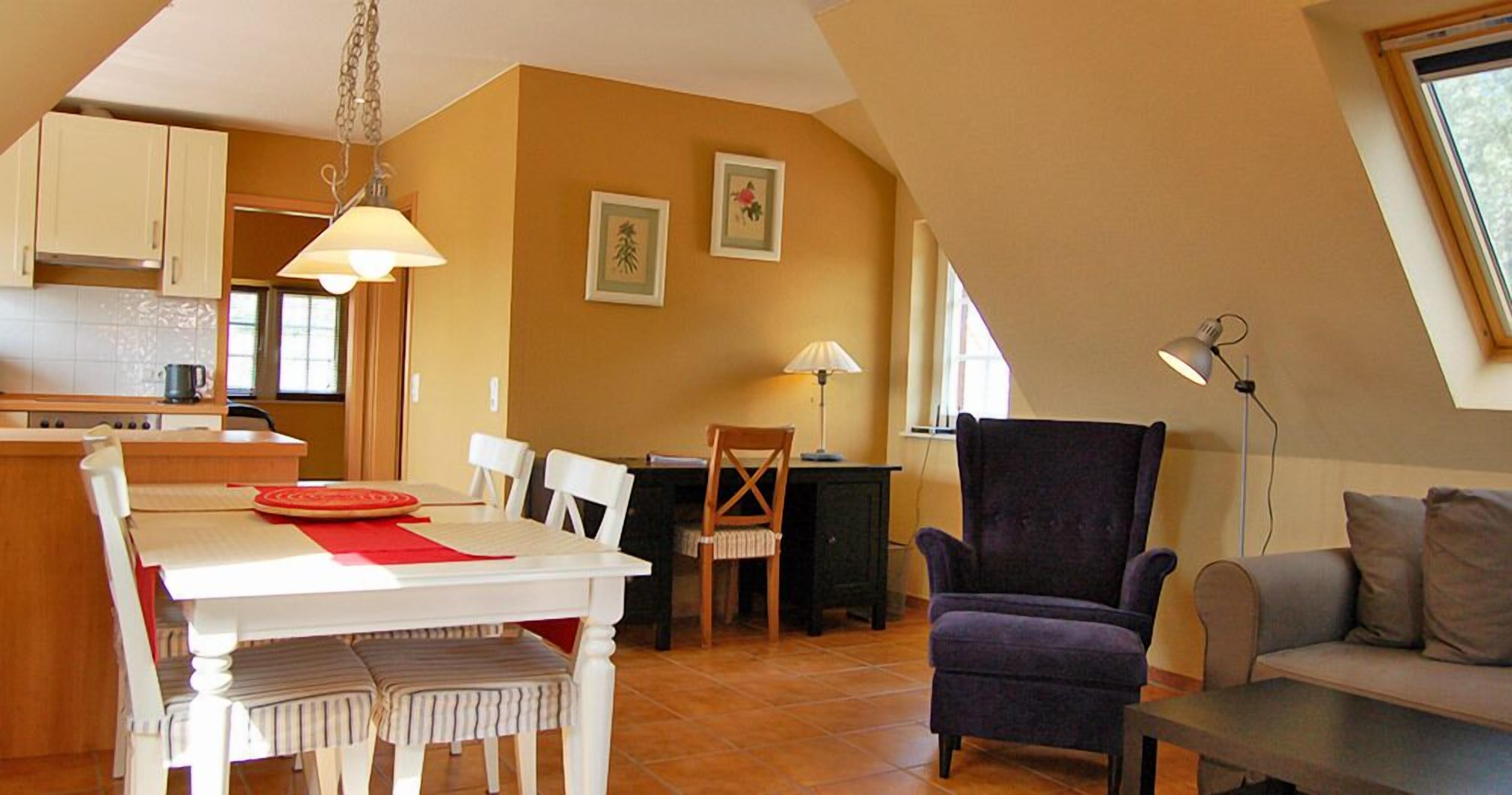 A cozy living room with a dining table, a purple armchair, and a sofa. There is a kitchen area with cabinets and a hanging light fixture.