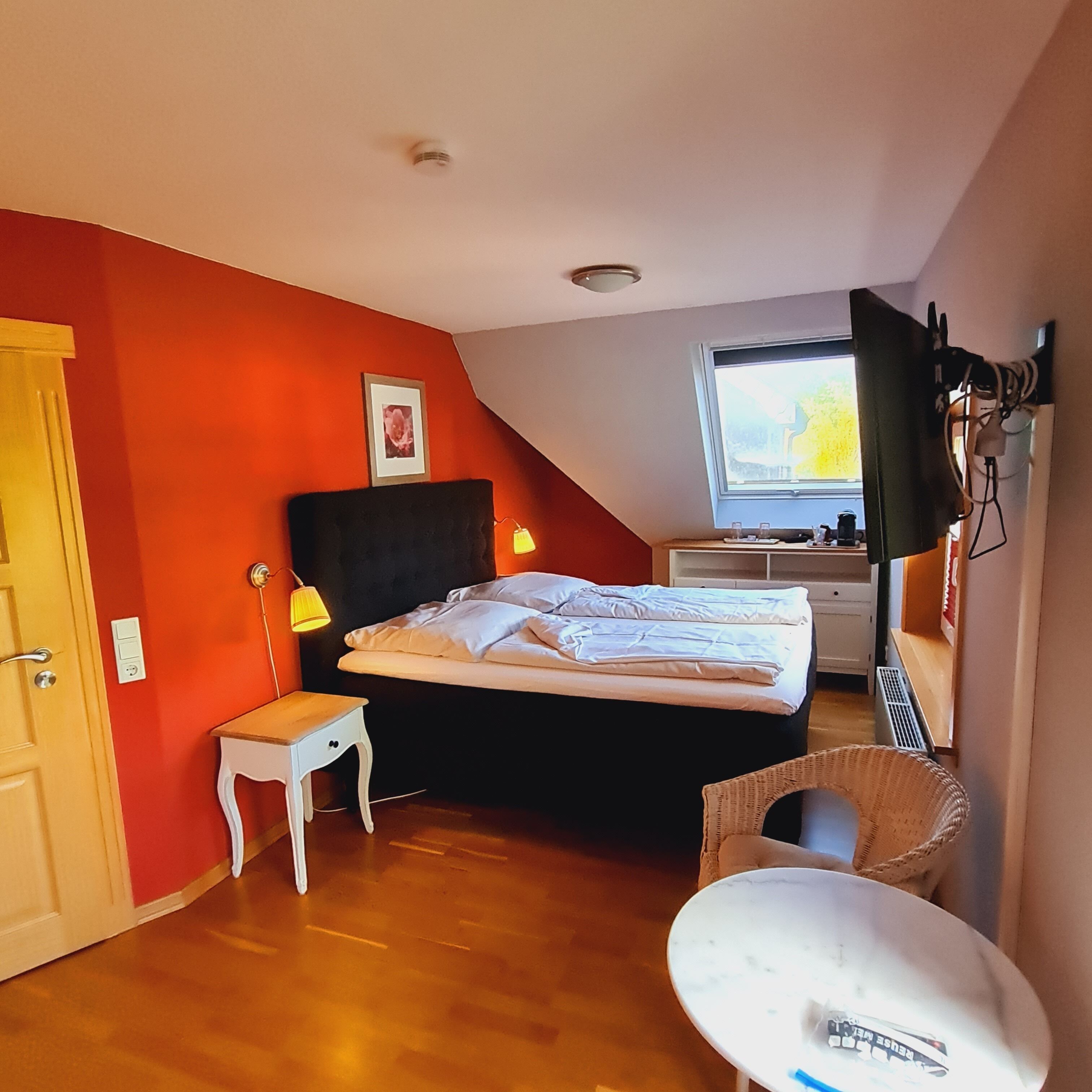 Ein Schlafzimmer mit einem Bett, einem kleinen Tisch, einem Stuhl, einem Fenster und einem an der Wand montierten Fernseher.