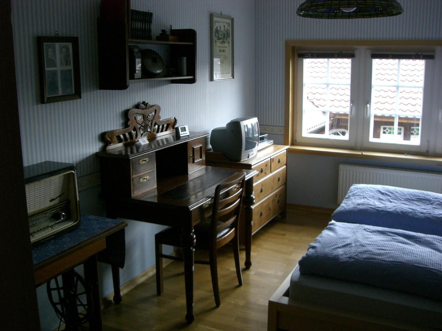 Et soveværelse med seng, skrivebord med computer, stol og et vindue med udsigt til bygninger.
