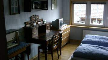 Et soveværelse med seng, skrivebord med computer, stol og et vindue med udsigt til bygninger.