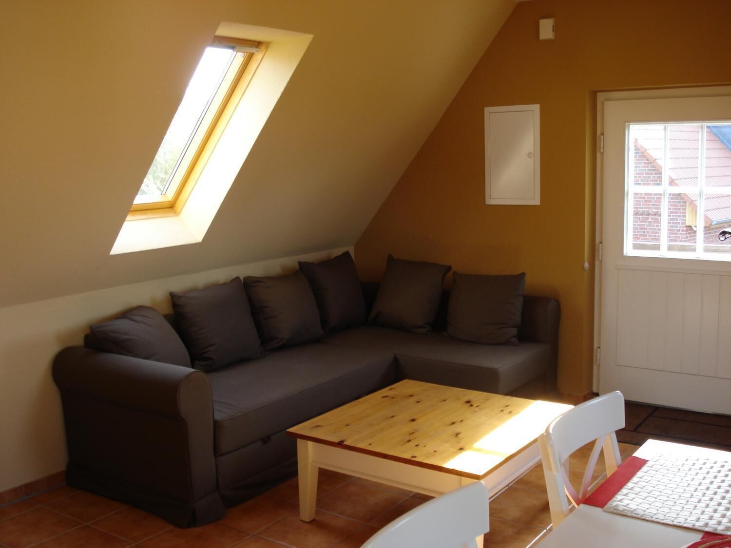 Ein gemütliches Zimmer mit einem braunen Sofa, einem Holztisch und einem weißen Stuhl. Ein Fenster bietet Blick auf ein Backsteingebäude.