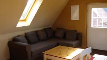 Ein gemütliches Zimmer mit einem braunen Sofa, einem Holztisch und einem weißen Stuhl. Ein Fenster bietet Blick auf ein Backsteingebäude.