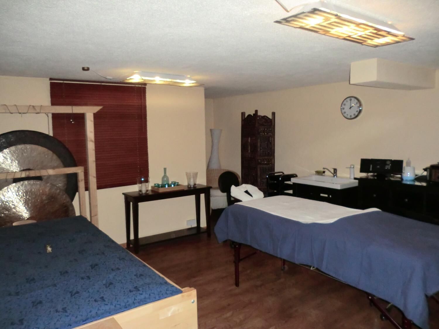 Ein Zimmer mit einem Bett, einem Schreibtisch, einem Stuhl, einer Wanduhr und einem Fenster mit Jalousien.