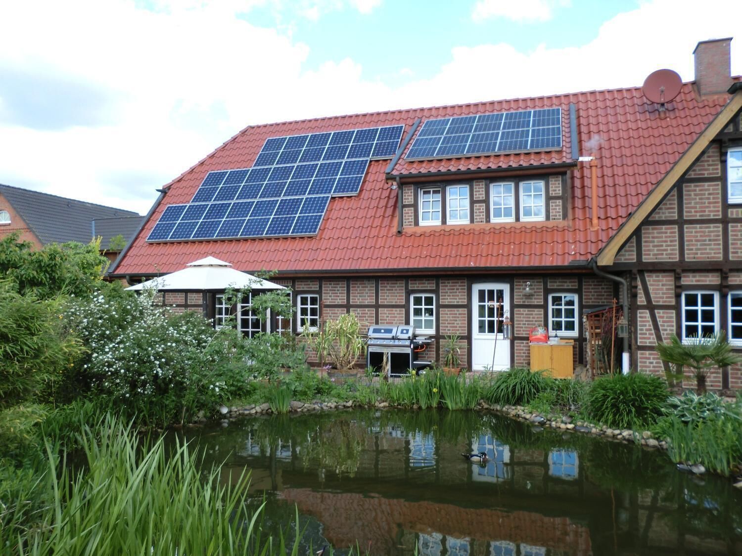 Una casa con paneles solares en el techo, un estanque y una zona de barbacoa.