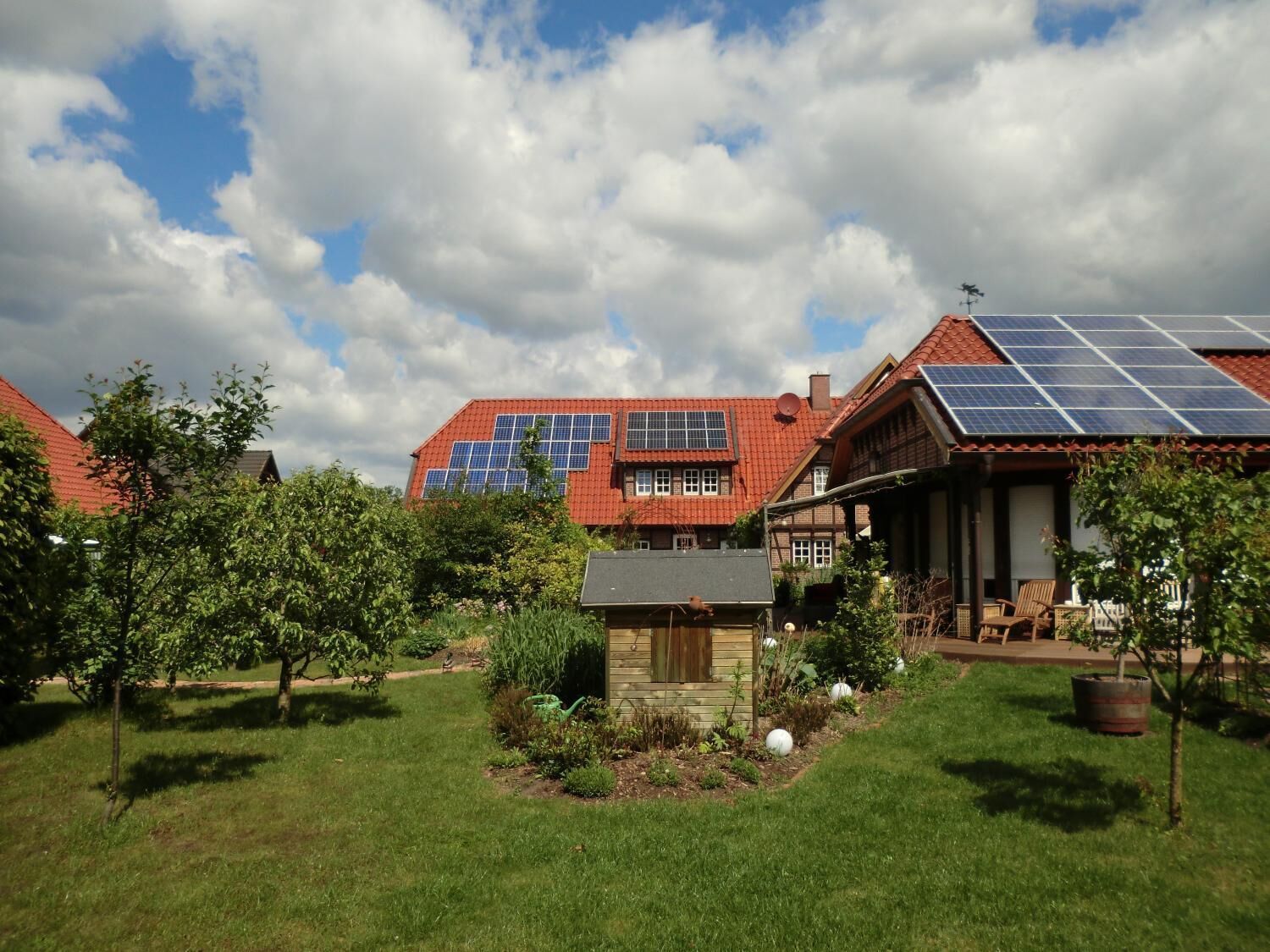 Een huis met zonnepanelen op het dak, omgeven door een verzorgde tuin en een kleinere houten constructie.