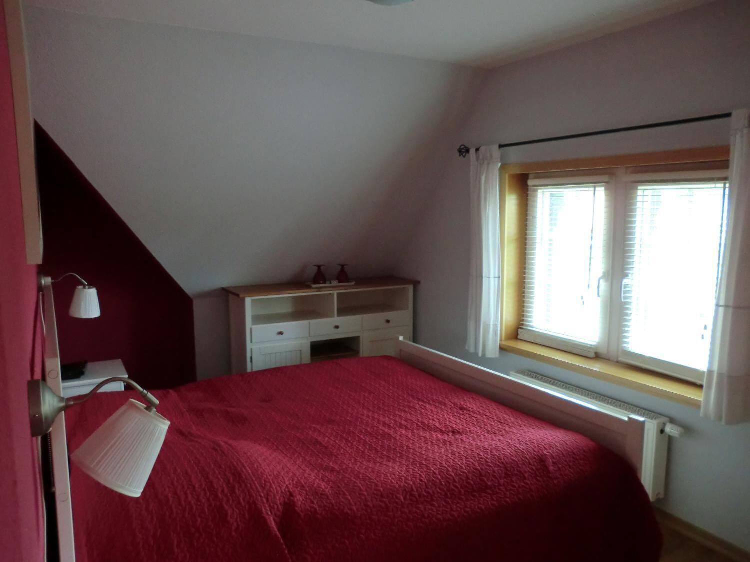 Ein Schlafzimmer mit schrägen Wänden, einem Bett mit roter Bettdecke, einem weißen Nachttisch mit zwei Lampen und einem Fenster mit Jalousien.