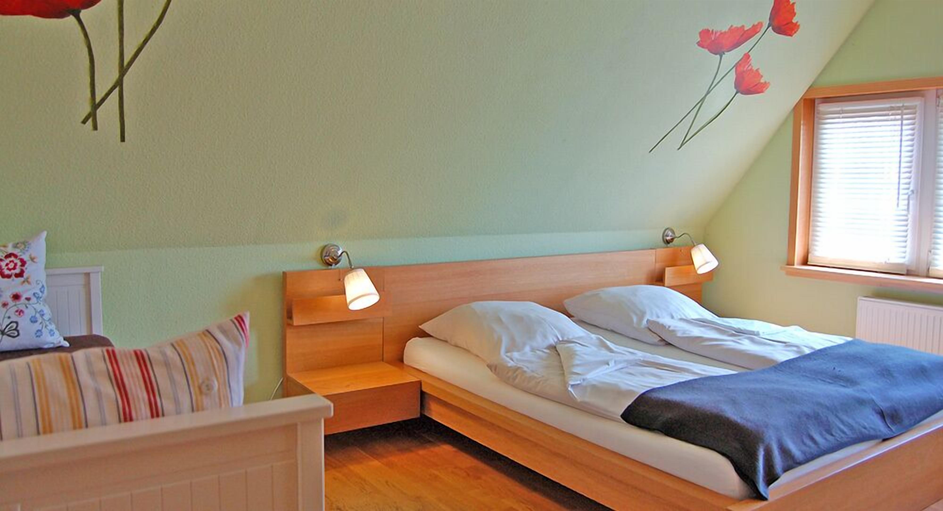 Ein Schlafzimmer mit einem Holzbett, zwei Nachttischlampen, einem Nachttisch, einem Fenster mit Jalousien und einer Wand mit floralen Verzierungen.