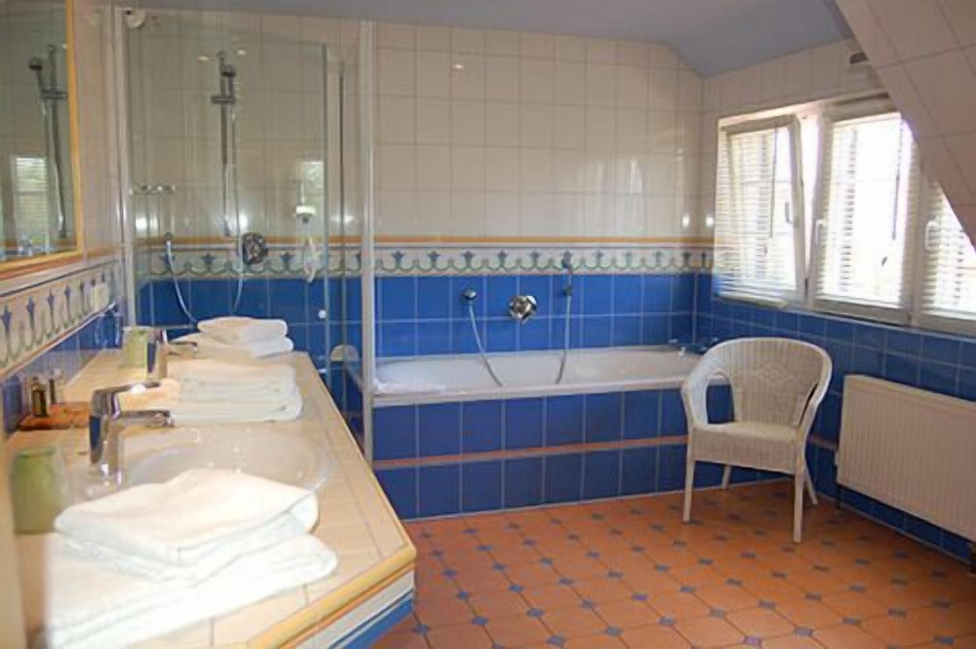 A bathroom with a bathtub, a shower, and a sink with a mirror. There is a chair and a window with blinds.
