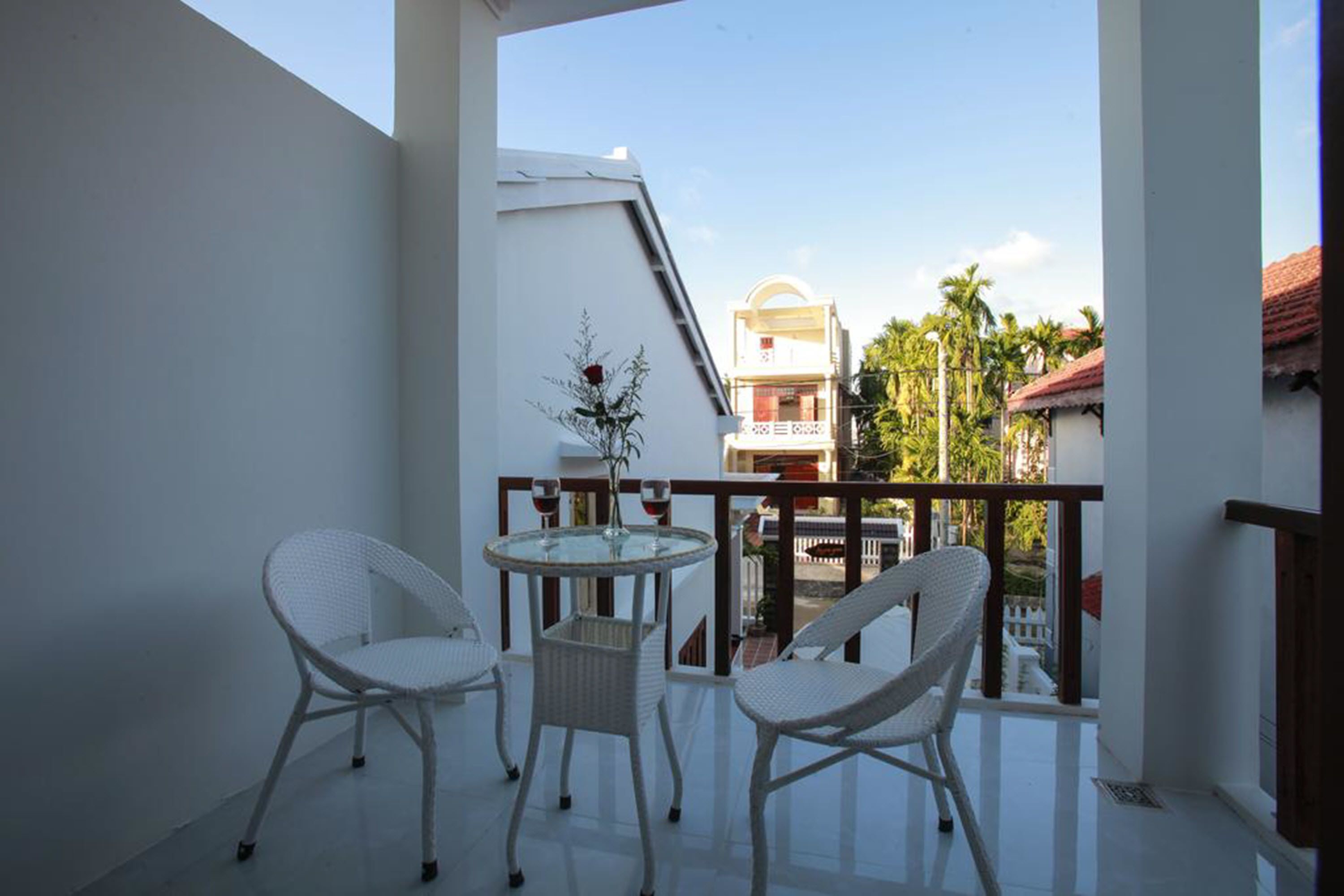 A balcony with white wicker chairs and a glass table, overlooking a residential area with palm trees and a building with a red roof.