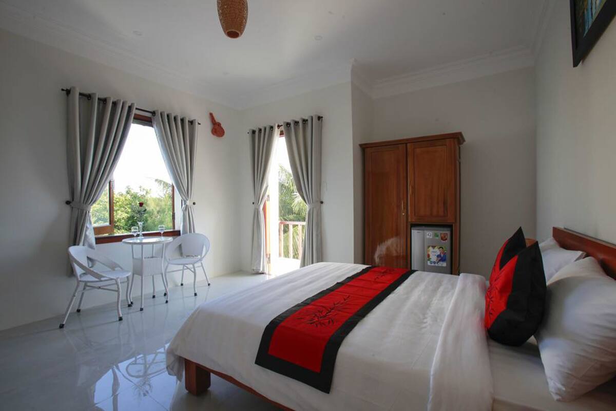 A bedroom with a bed, a wooden wardrobe, a small table with chairs, and a window with curtains.