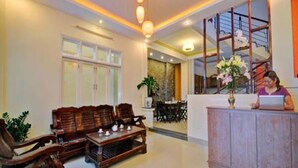 A reception area with a wooden reception desk, a sofa set, and a dining area in the background. - Coconut Garden Homestay Hoi An (Da Nang)