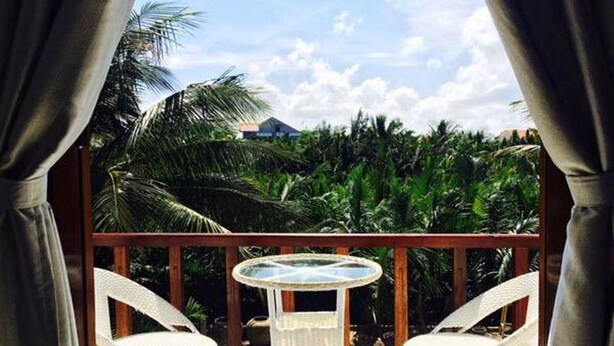 A balcony with two white wicker chairs and a small round table, overlooking a lush green landscape with palm trees and a building in the background. - Coconut Garden Homestay Hoi An (Da Nang)