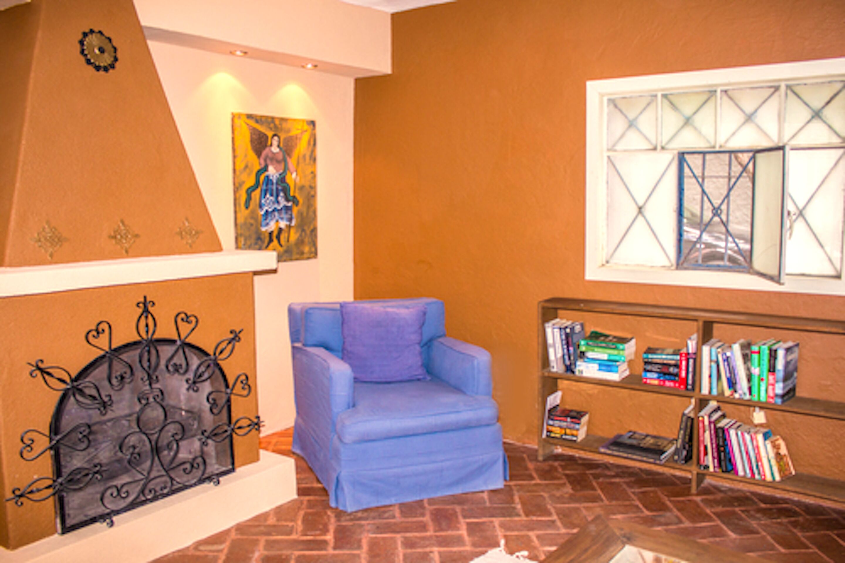 A room with a blue armchair, a bookshelf with books, a painting on the wall, and a decorative metal headboard.